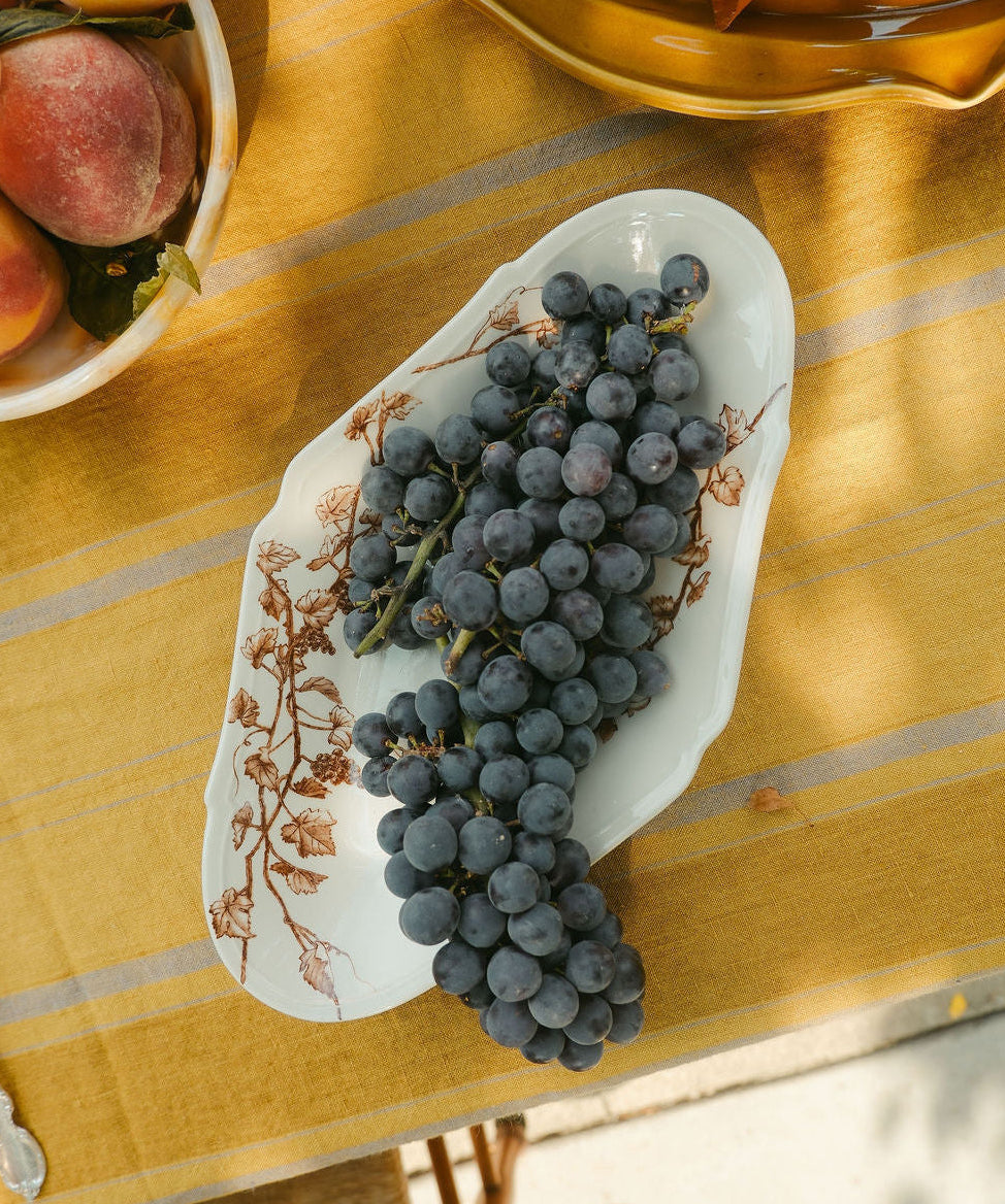 A white ceramic serving platter from Japan with brown floral tissue transfer vine patterns, filled with dark purple grapes, set on a yellow tablecloth with a rustic outdoor dining scene.
