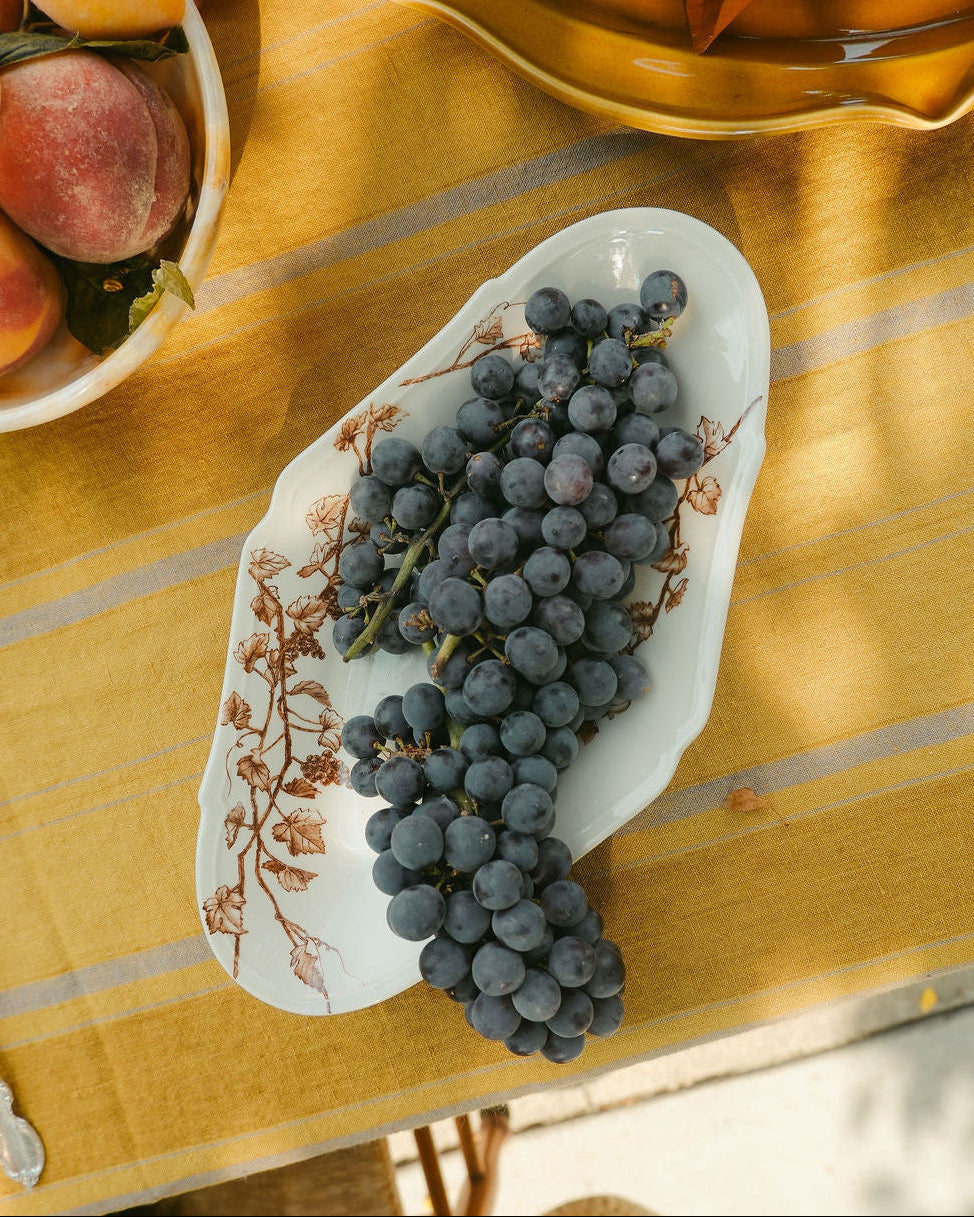 A white ceramic serving platter from Japan with brown floral tissue transfer vine patterns, filled with dark purple grapes, set on a yellow tablecloth with a rustic outdoor dining scene.