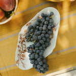 A white ceramic serving platter from Japan with brown floral tissue transfer vine patterns, filled with dark purple grapes, set on a yellow tablecloth with a rustic outdoor dining scene.