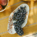 A white ceramic serving platter from Japan with brown floral tissue transfer vine patterns, filled with dark purple grapes, set on a yellow tablecloth with a rustic outdoor dining scene.